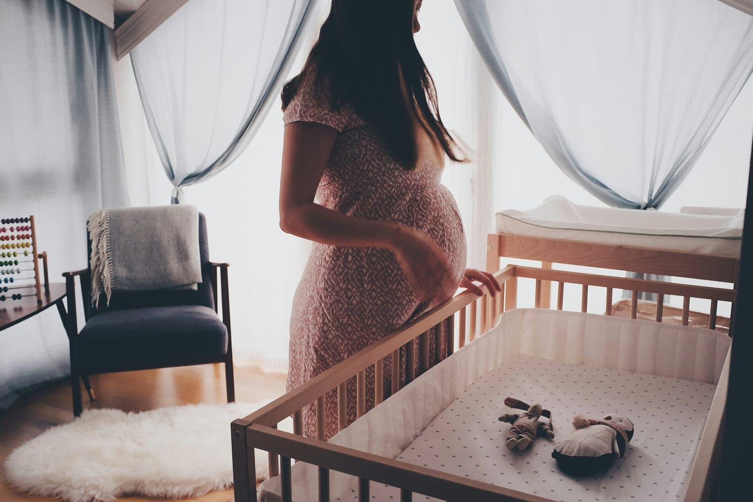 Expectant mother standing beside a wooden crib in a bright, sunlit nursery