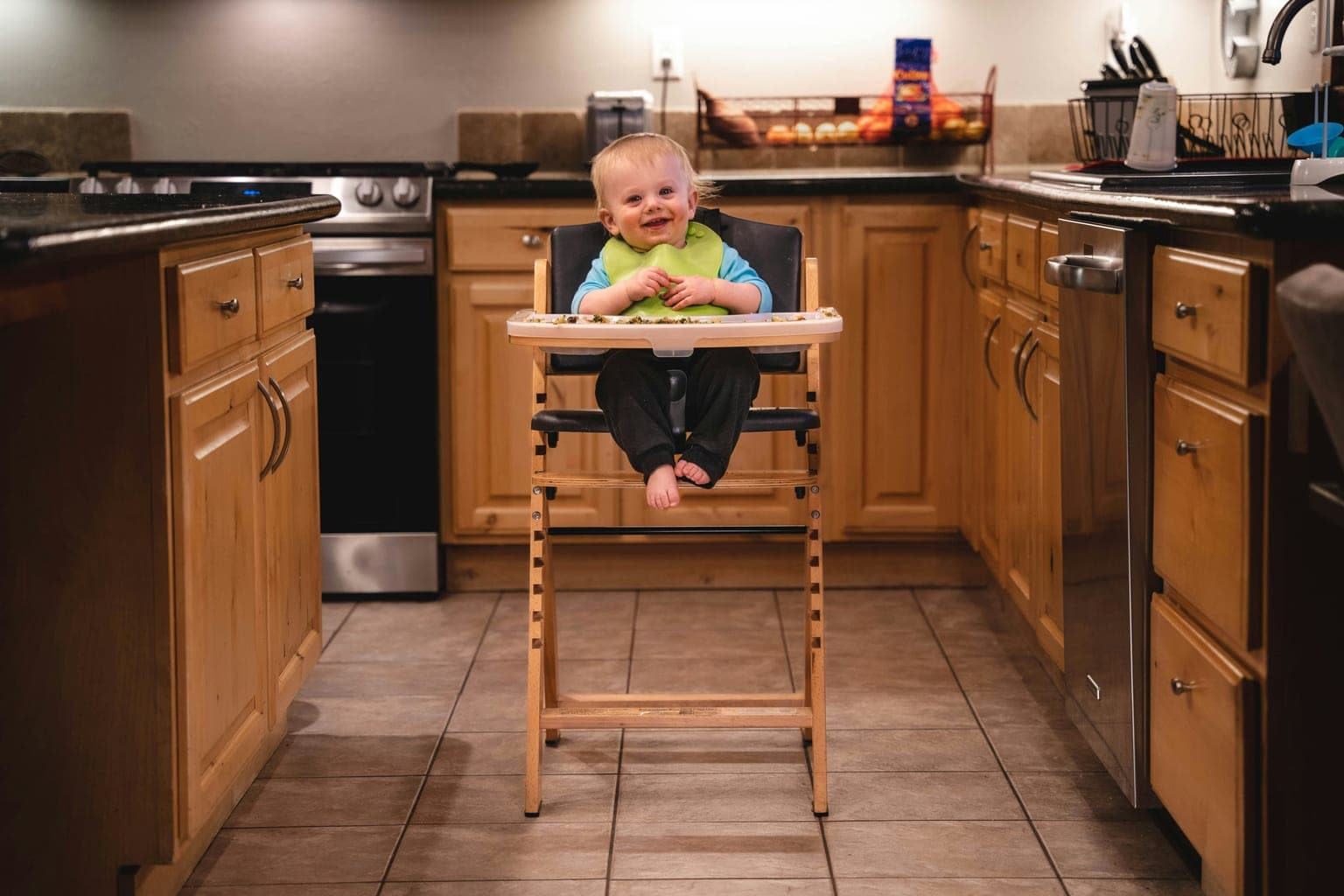 Toddler in a green bib smiling while sitting in a wooden high chair in a kitchen