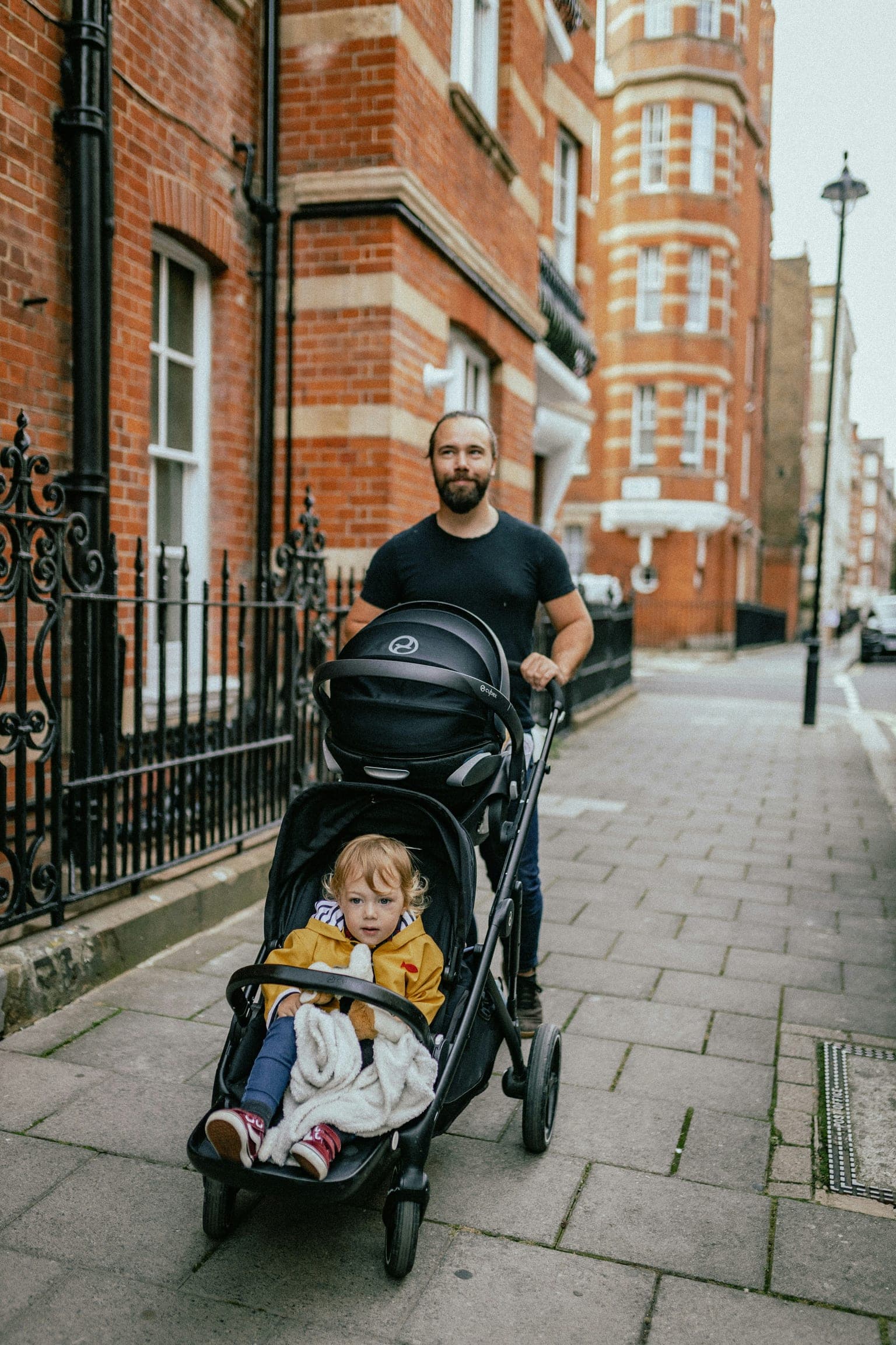 Father pushing a stroller with a toddler in a yellow raincoat along a city sidewalk